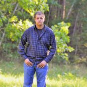 A young man stands in a forest wearing a plaid shirt and denim jeans.